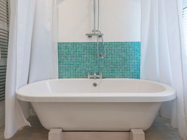 A white bathtub with a silver faucet and showerhead against a wall with small green tiles and white curtains at Whitsunday House in Carbis Bay