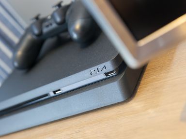 A close-up of a black PS4 console with a controller on top next to a television on a wooden surface