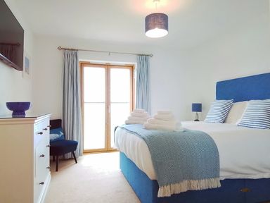 A bedroom with a blue bed and striped pillows a dresser and chair near a window at Sea Mist in Carbis Bay