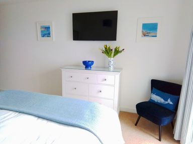 A bedroom with a white dresser holding a blue bowl and a vase with tulips a wall-mounted TV two framed pictures a blue chair with a whale cushion and a bed with a light blue blanket at Sea Mist in Carbis Bay