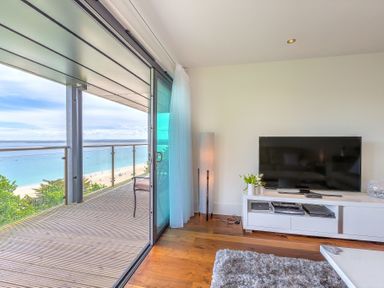 A living room with a television on a cabinet and sliding glass doors opening to a balcony with ocean view at Abalone in St Ives