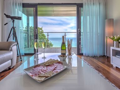 A living room with a plate of salami and crackers on a table with champagne and a telescope by a window overlooking the sea at Abalone in St Ives