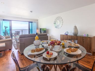 A dining table set for breakfast with croissants fruit and tea in a living room with sofas and a balcony overlooking the sea at Abalone in St Ives