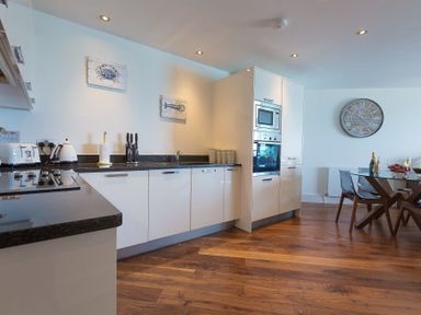 A kitchen with white cabinets a black countertop a built in microwave oven and a glass dining table with chairs at Abalone in St Ives