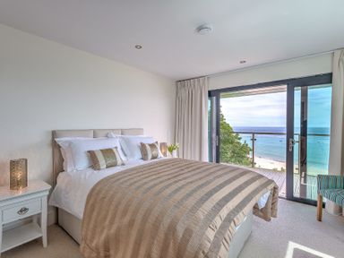A bedroom with a large bed, bedside table with lamp, sliding glass doors opening to a balcony with a sea view at Abalone in St Ives