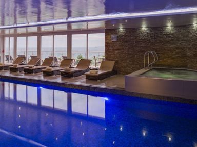 An indoor swimming pool with lounge chairs and a hot tub near large windows overlooking the sea at Abalone in St Ives