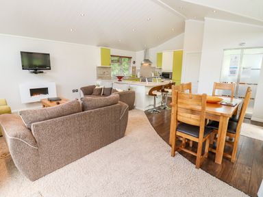 An open living space with brown sofas a wooden dining table with chairs a kitchen island with bar stools and a wall mounted television at Oak Lodge in Glasshouse