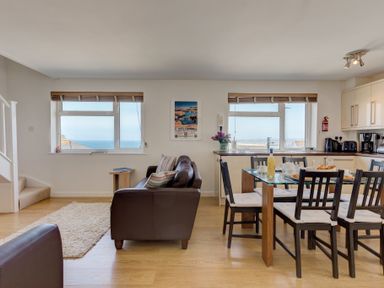 A living room and kitchen with a brown sofa wooden chairs and a glass dining table at Atlantic Blue in Carbis Bay