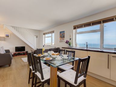 A dining area with a glass table set for breakfast and a living room with sofas and a television near a staircase at Atlantic Blue in Carbis Bay