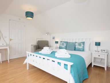 A bedroom with a white bed and turquoise bedding with pillows and white towels on the bed with a striped armchair and white bedside tables at Atlantic Blue in Carbis Bay