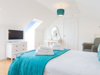 A bedroom with a bed covered in white sheets and turquoise blanket with folded towels a dresser with a TV a vanity table with a mirror a striped armchair and a skylight window at Atlantic Blue in Carbis Bay