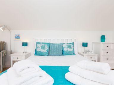 A bedroom with a white bed featuring blue and turquoise pillows and bedding with white towels on the bed and two white bedside tables with lamps and a striped armchair at Atlantic Blue in Carbis Bay