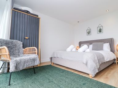 A bedroom with a bed with folded towels on it a gray armchair a dark wardrobe and botanical prints on the wall at Atlantic Watch 8 in Carbis Bay