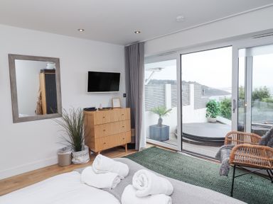 A bedroom with a bed towels on it a dresser a wall mirror a plant a wall mounted tv and sliding glass doors leading to a balcony with a hot tub and two plants at Atlantic Watch 8 in Carbis Bay