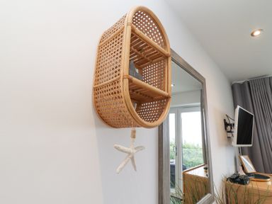 A rattan wall-mounted shelf with a hanging starfish decoration and a mirror reflecting a window and a television at Atlantic Watch 8 in Carbis Bay