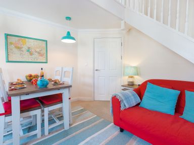 A dining area with a wooden table and white chairs with red cushions and a red sofa with blue cushions under a staircase at Brinyloft in Carbis Bay