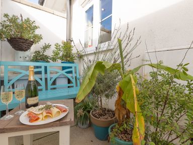 A small outdoor patio with a wooden table holding a plate of cheese and meat and two glasses of sparkling wine with a blue bench and several potted plants at Brinyloft in Carbis Bay