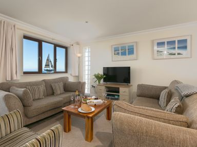 A living room with two sofas a TV on a stand a wooden coffee table with food and drinks and a window with a sailboat decoration at Cornerways in Carbis Bay
