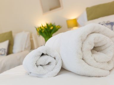 Rolled white towels on a bed with a lamp and flowers in the background at Cornerways in Carbis Bay