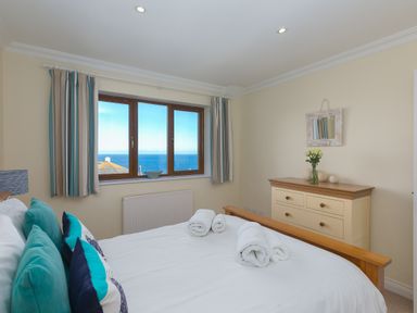 A bedroom with a bed pillows and rolled towels by a window showing the sea and a dresser with flowers at Cornerways in Carbis Bay