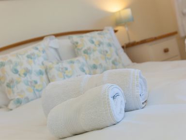 Two rolled white towels on a bed with floral pillows and a lamp in a bedroom at Cornerways in Carbis Bay