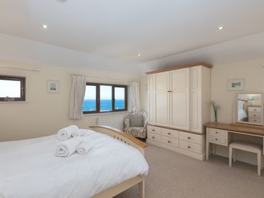 A bedroom with a bed, rolled towels on the bed, an armchair, a wardrobe, a dressing table with a stool, and a window with a sea view at Cornerways in Carbis Bay