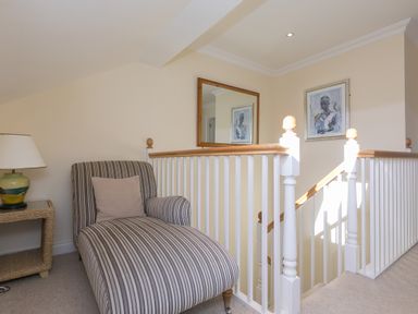 A striped chaise lounge chair next to a wicker side table with a lamp in a hallway by a staircase with white railings and a mirror on the wall at Cornerways in Carbis Bay
