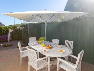 An outdoor patio area with a wooden table and six chairs under a large white umbrella with plates of fruit and croissants at Cornerways in Carbis Bay