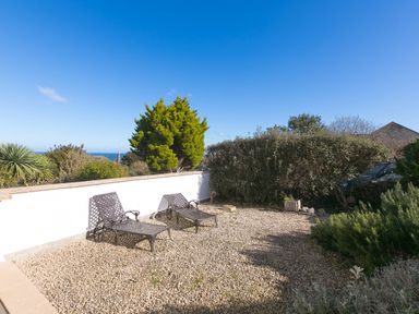 An outdoor area with two metal lounge chairs on a gravel surface a white wall hedges and trees in the background at Cornerways in Carbis Bay