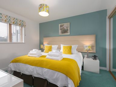 A bedroom with a double bed yellow cushions and blanket bedside tables with lamps a window with blinds and a picture on the wall at Elanora in Carbis Bay