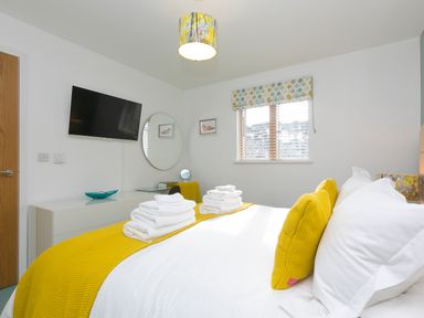 A bedroom with a bed with yellow pillows and blanket stacked white towels a wall mounted TV a round mirror and a window with blinds at Elanora in Carbis Bay