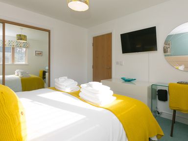 A bedroom with a bed covered in white linens and yellow blankets and towels a wall-mounted tv a wooden door a mirrored wardrobe and a glass desk with a yellow chair at Elanora in Carbis Bay