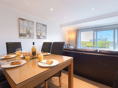 A dining table set with glasses of orange juice and pastries next to a leather sofa in a room with a floor lamp and sliding glass doors at Gennaker in Carbis Bay