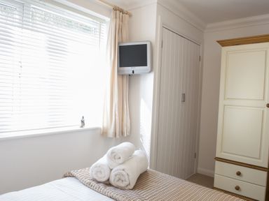 A bedroom with a window with blinds and curtains a small wall-mounted television a wardrobe a closet and rolled towels on the bed at Gennaker in Carbis Bay