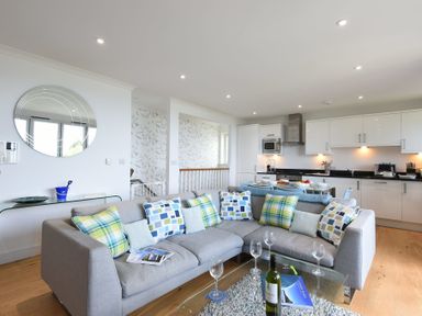 A living room with a grey sectional sofa and kitchen with white cabinets in the background at Halcyon Cove in Carbis Bay