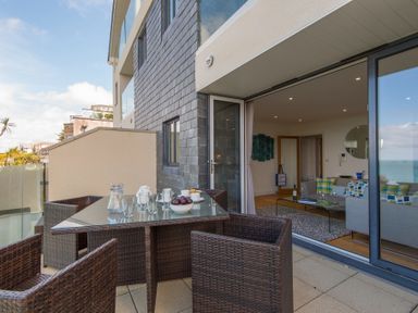 An outdoor patio with a glass table and wicker chairs next to a living room with sofas and a coffee table at Halcyon Cove in Carbis Bay