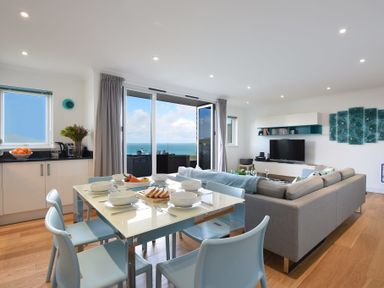 A dining table set with bowls and cutlery next to a living room with a sofa and TV in a room with sliding doors overlooking the sea at Halcyon Cove in Carbis Bay