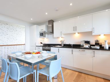 A kitchen with white cabinets black countertops a dining table set with blue chairs and dishes at Halcyon Cove in Carbis Bay