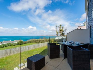 An outdoor patio with a glass table and chairs overlooking the ocean at Halcyon Cove in Carbis Bay