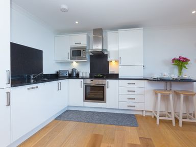 A kitchen with white cabinets black countertops a microwave oven a toaster a kettle and stools at Kernow's Dream in Carbis Bay