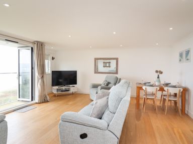 A living room with grey upholstered chairs a wooden dining table with chairs and a television at Kernow's Dream in Carbis Bay
