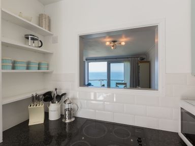 A kitchen corner with a black countertop knives and utensils on the counter and an open window showing a room and ocean view at Ocean Cove in Carbis Bay