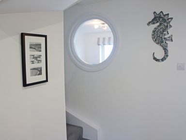 A hallway with a framed picture on the wall a decorative seahorse hanging and a round window showing another room at Ocean Cove in Carbis Bay