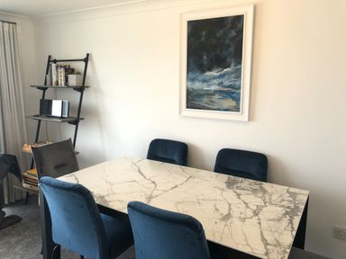 A dining room with a marble table and blue chairs a ladder shelf with books and a framed painting on the wall at Ocean Cove in Carbis Bay