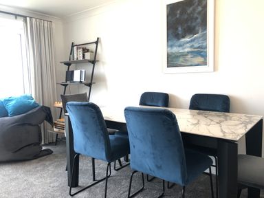 A dining room with a marble table and blue chairs a gray armchair with blue cushions a black leaning shelf with books and a stereo and a landscape painting on the wall at Ocean Cove in Carbis Bay