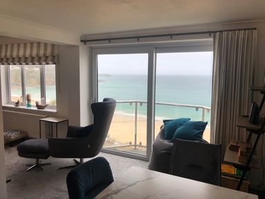 A living room with an armchair and ottoman near large windows overlooking a beach at Ocean Cove in Carbis Bay