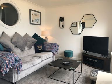 A living room with a gray sofa and cushions a black metal coffee table a television on a wooden stand and decorative wall mirrors at Ocean Cove in Carbis Bay