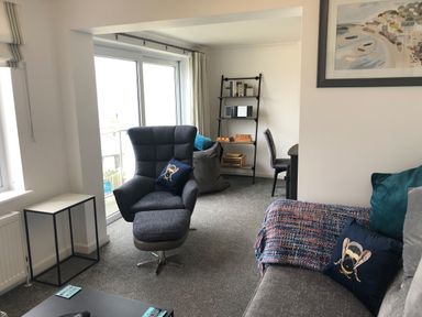 A living room with a grey armchair with a footrest blue cushions a grey sofa with cushions a coffee table a side table a bookshelf and a chair at Ocean Cove in Carbis Bay