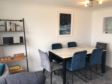 A dining area with a marble top table and blue and grey chairs with a painting on the wall and a bookshelf with books and a stereo at Ocean Cove in Carbis Bay