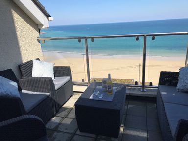 A balcony with wicker chairs a table and drinks overlooking a beach and ocean at Ocean Cove in Carbis Bay